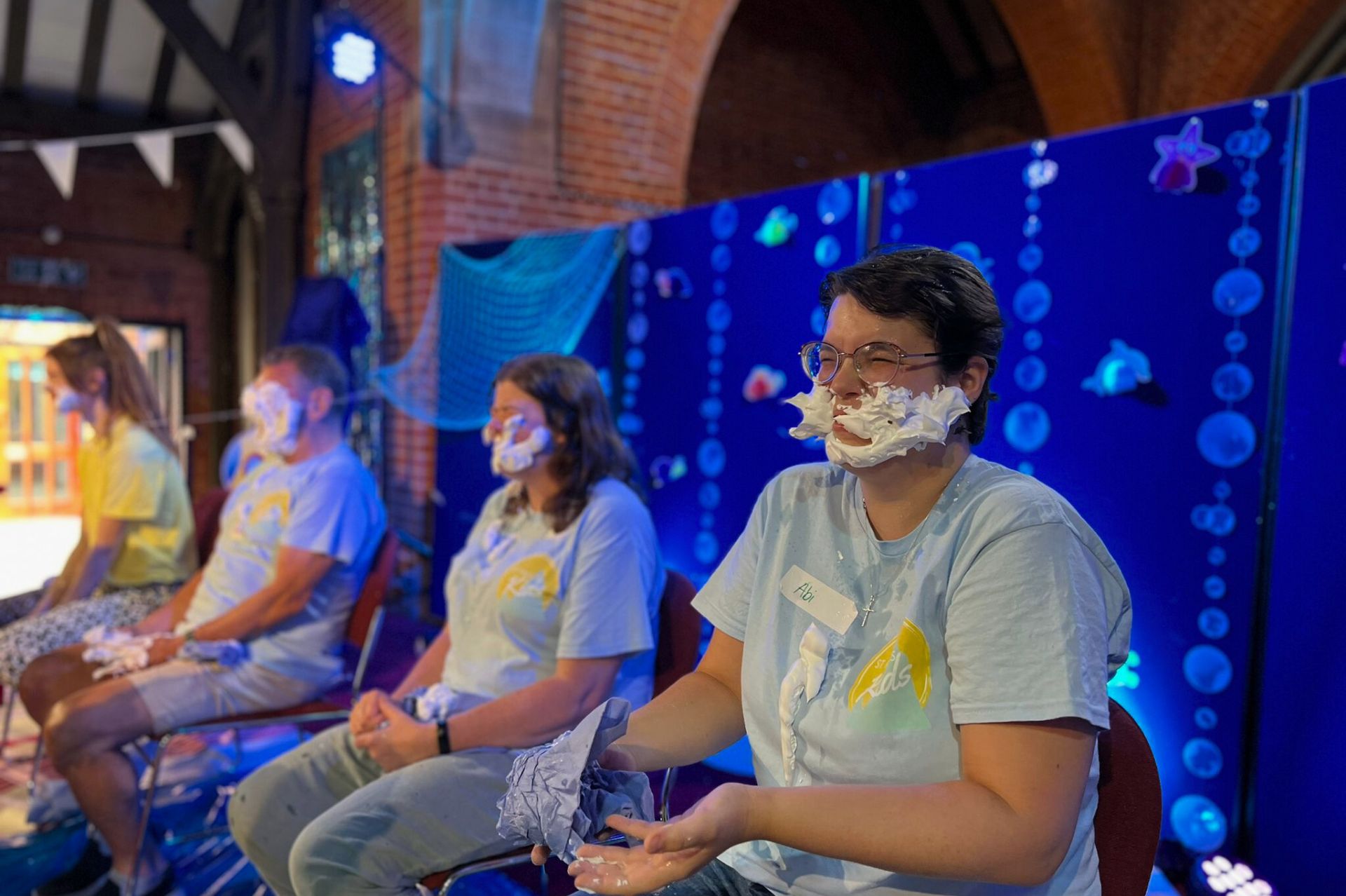 A group of adults in holiday club t-shirts sat on chairs with their faces covered by shaving foam