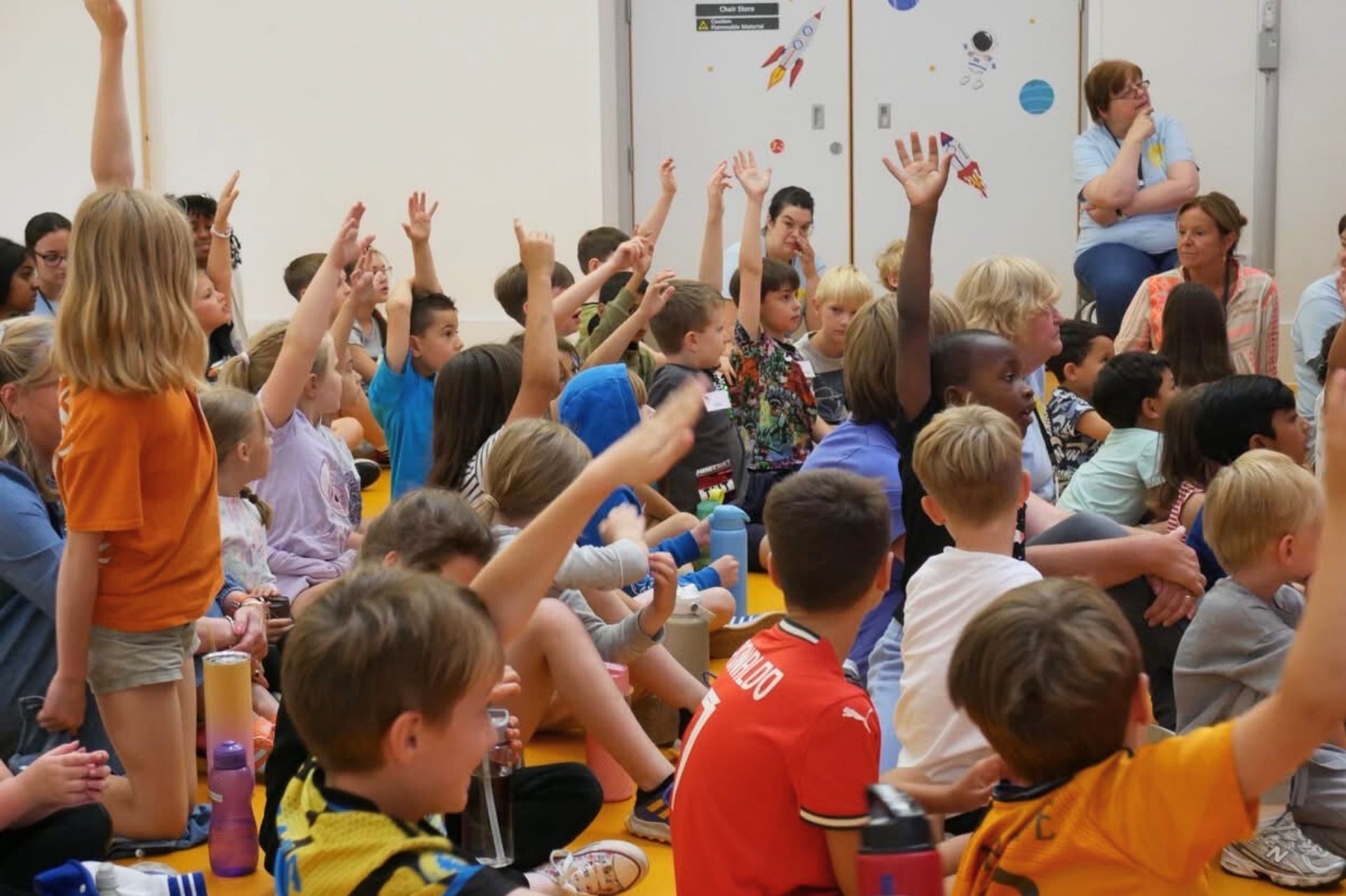 A large number of children sat on a hall floor excitedly putting their hand up