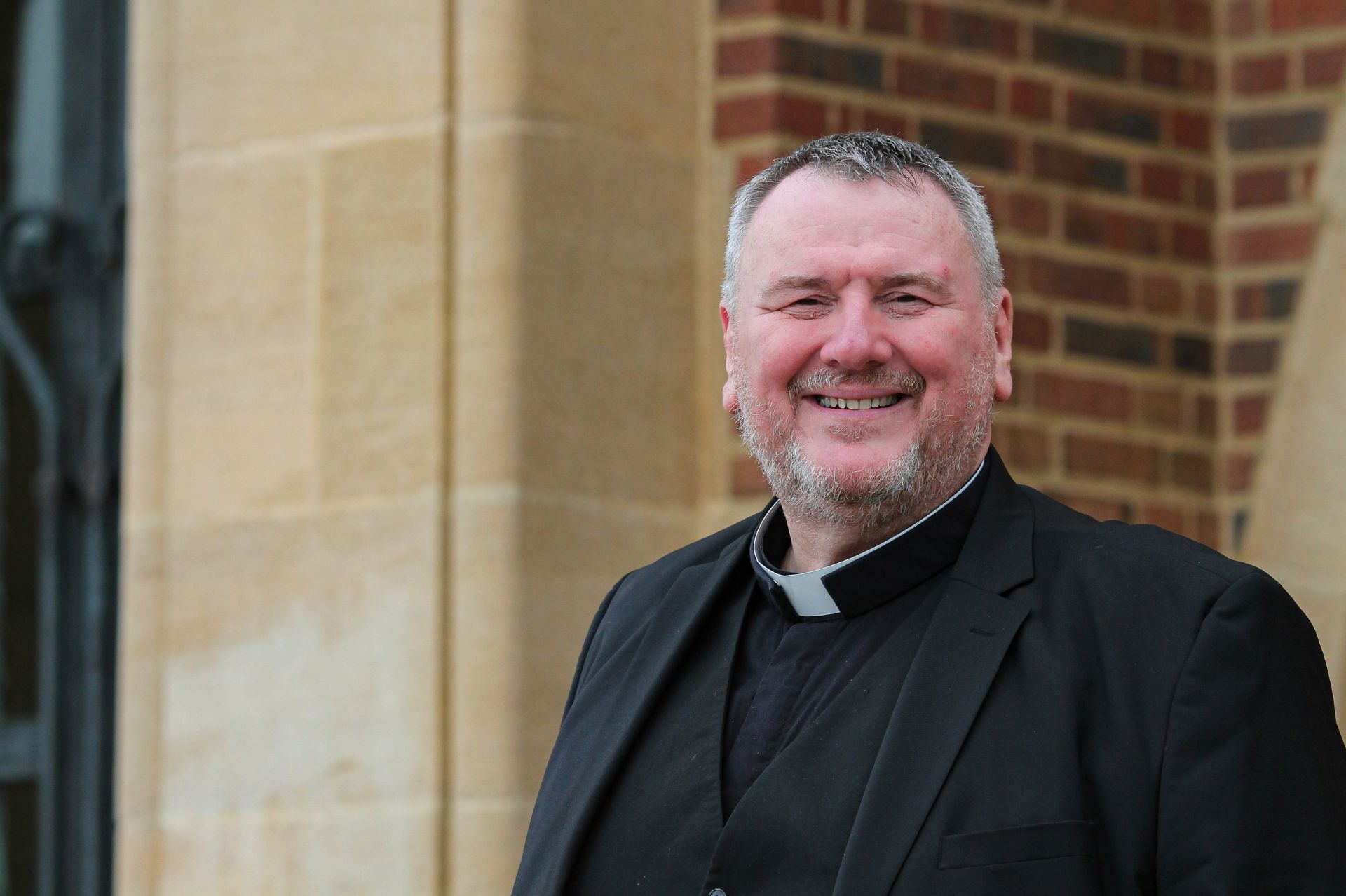 The Very Revernd Bob Cooper outside Guildford Cathedral wearing a black clerical shirt, waistcoat and jacket