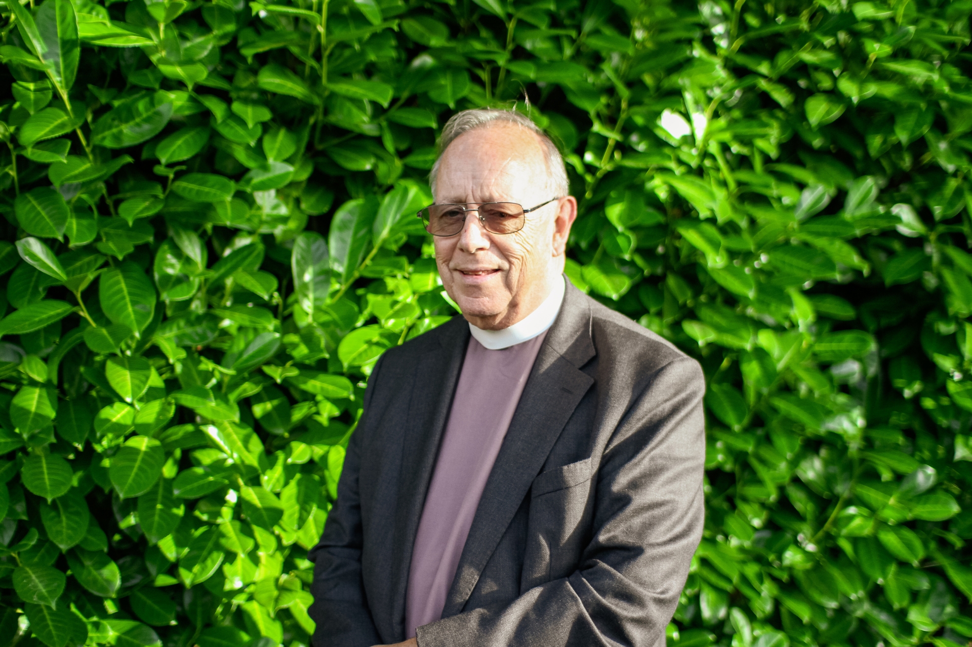 The Venerable Stuart Beake stood in front of a green hedge wearing a pastel purple clerical shirt and grey jacket