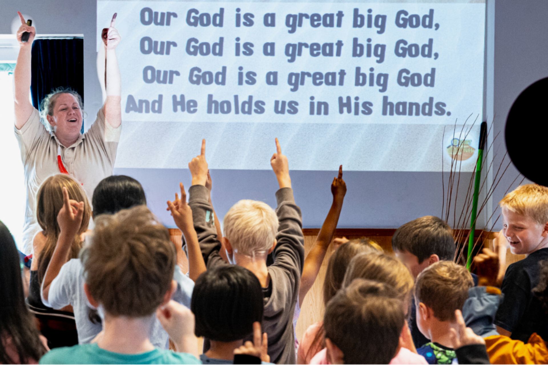 Woman leading action songs next to a projector screen and in front of a group of children who are joining in