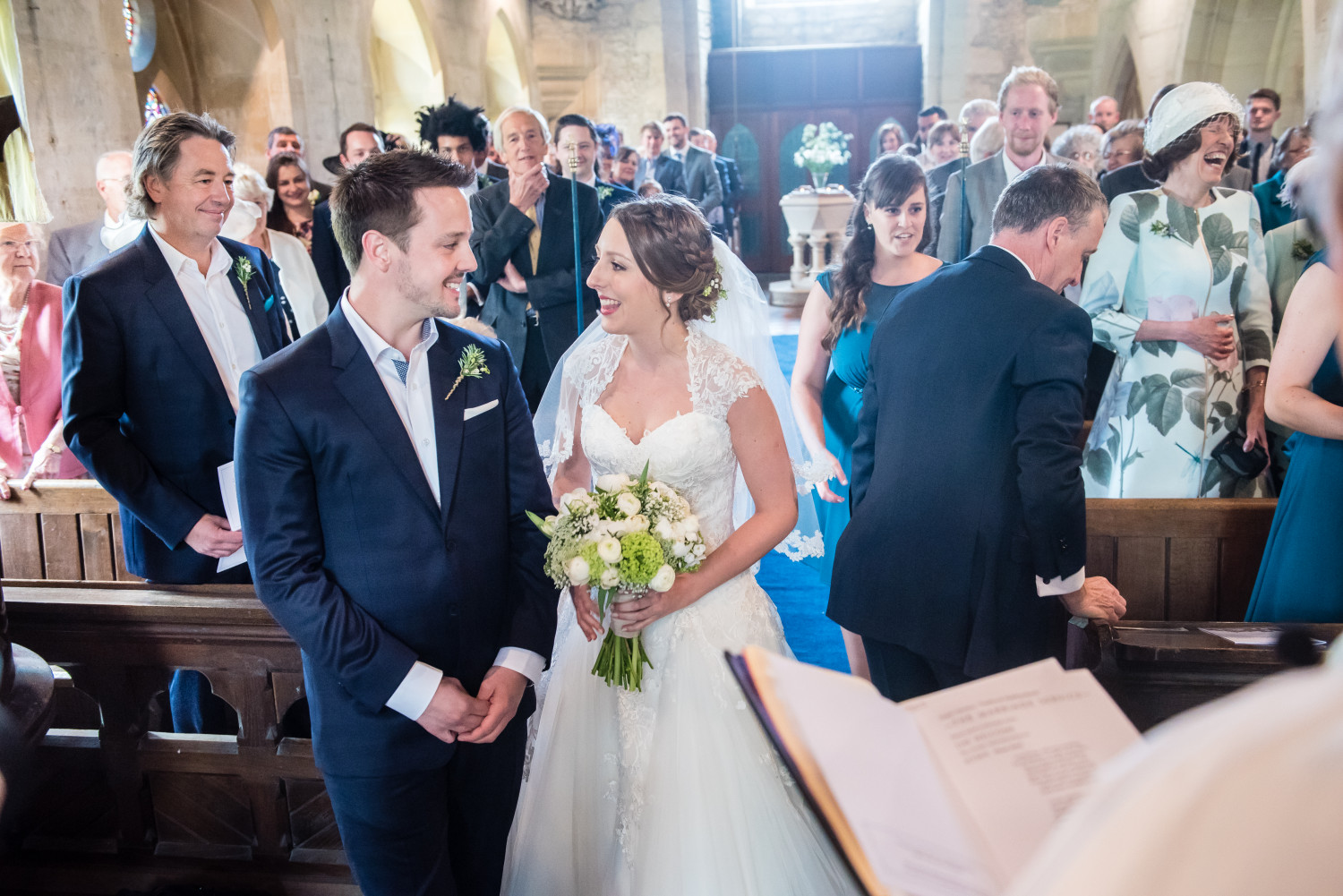 A man in a smart navy suit and a woman in a white wedding dress getting married, whilst their friends and family watch on