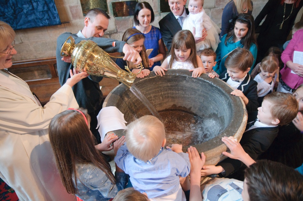 Priest pours water from an ornate polished brass jug into a baptism font, surrounded by parents and children. 