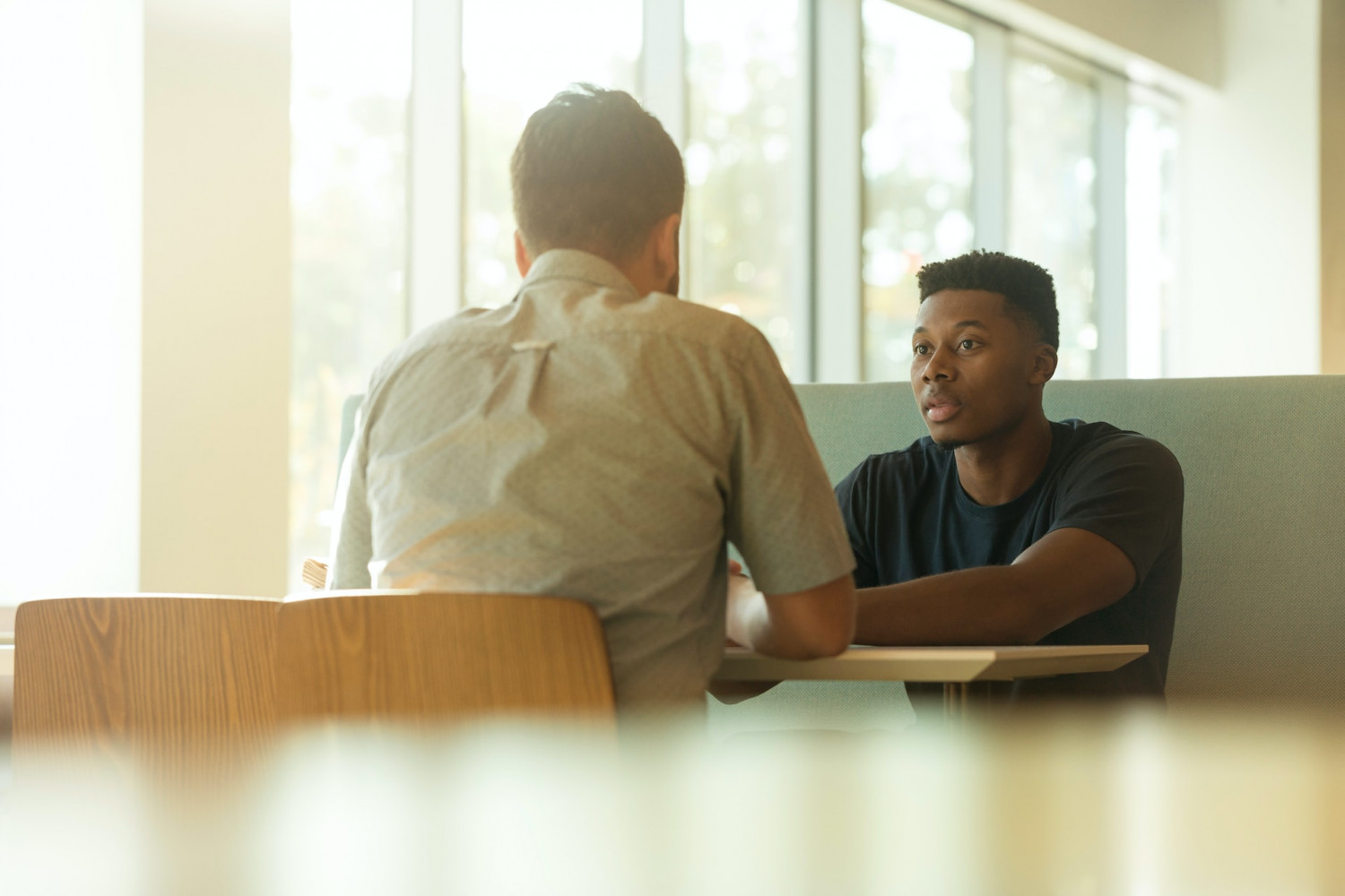 Two young men sat at the table facing each other deep in conversation in a sunlit room